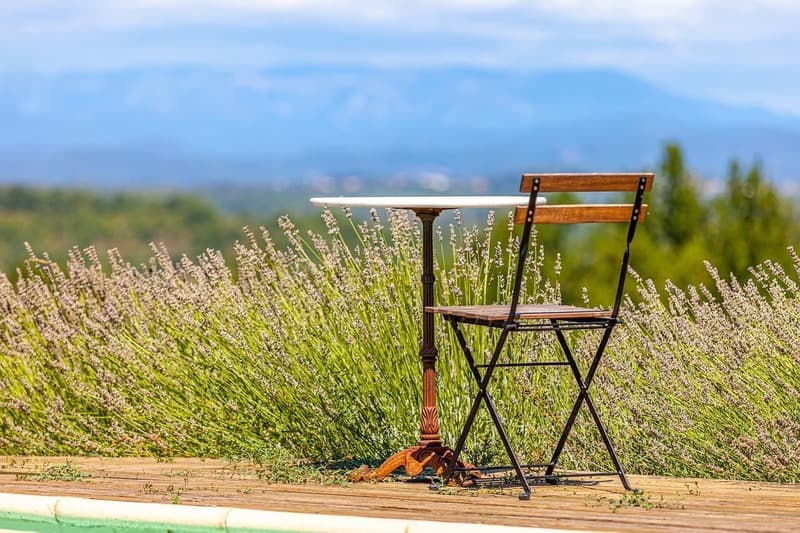 transmission-cabinet-expertise-comptable-VG Chaise et table de bistrot en bois et métal, posées sur une terrasse en bois devant un champ de lavande ou de fleurs et une chaîne de montagnes floue en arrière-plan.