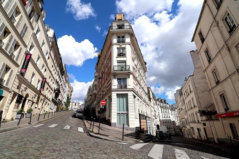 Immeuble d'angle à l'architecture typique de Paris ou d'une grande ville française, au croisement d'une rue pavée montante sous un ciel bleu nuageux.