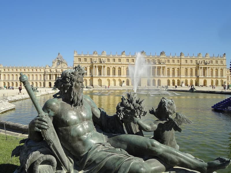 Statue en bronze de Neptune avec une allégorie, jet d'eau dans le Bassin de Neptune, et le Château de Versailles en arrière-plan sous un ciel bleu.