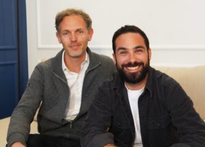 Laurent Charrier et Alexis SLAMA assis sur un canapé beige devant un mur blanc avec une porte bleue. Ils portent des vêtements décontractés et sourient à la caméra.