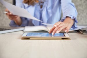 Personne en chemise bleue utilisant une tablette tout en consultant des documents papier sur un bureau en bois clair.