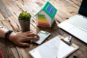 Calcul du bilan énergétique d'une maison avec maquette d'habitation à panneau solaire et étiquette énergétique, plante succulente et ordinateur portable sur bureau en bois.
