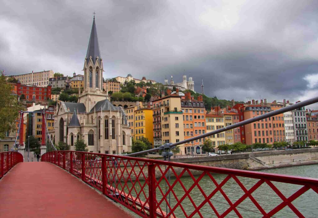 Église gothique, pont rouge, façades colorées, colline verte, ciel gris.