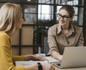 Deux femmes discutent lors d’une réunion professionnelle autour d’un ordinateur portable, dans un bureau moderne et lumineux.